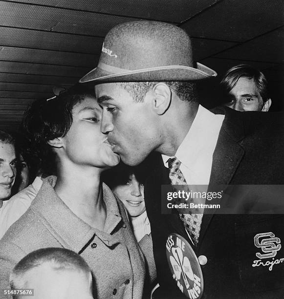 Wearing a button reading "Kiss Me, I'm Irish," University of Southern California football hero O.J. Simpson gets a welcome home kiss from his wife...