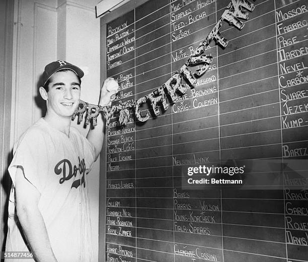 Dodgers baseball player Sandy Koufax at age 18 standing in front of a blackboard at Christmas wearing a baseball uniform.