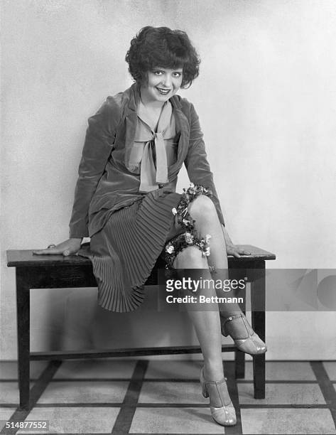 Clara Bow sitting on piano bench showing her flowered garters. Undated photograph.