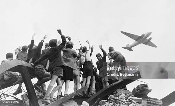 Group of German children stand atop building rubble, cheering a United States cargo airplane as it flies over a western section of Berlin. American...