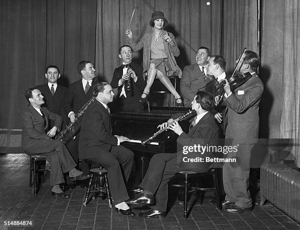 Smiling female bandleader conducts for her swing band as she sits atop a piano.