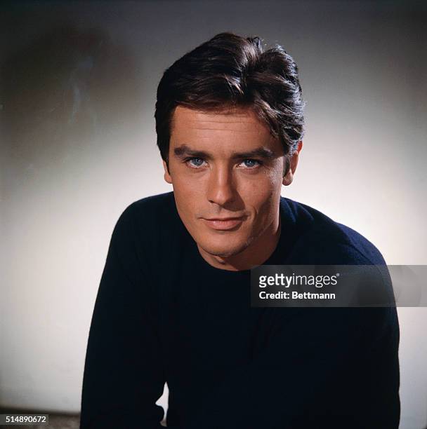 Head and shoulders portrait photo of French actor Alain Delon smiling in a black sweater. Ca. 1960s.