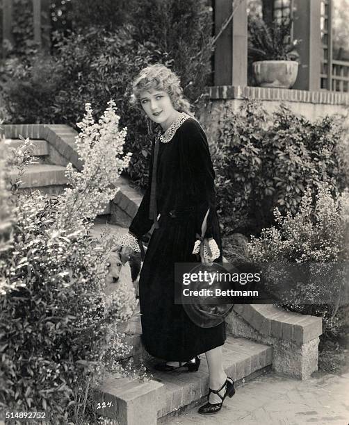 Mary Pickford, movie personality, standing on steps with a dog. She is wearing a velvet dress, designed by Jeanne Lanvin.