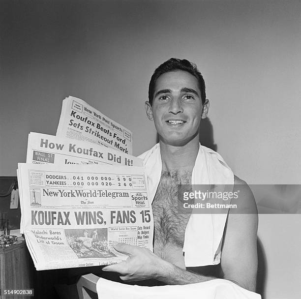 New York, NY: A big grin on his face, LA Dodgers star Sandy Koufax relaxes after bath in his Roosevelt Hotel Suite, holding newspapers which headline...
