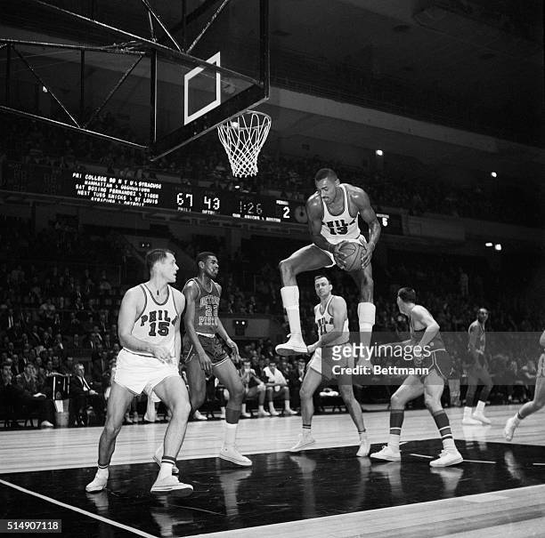 New York, NY- High and mighty is Philadelphia's Wilt Chamberlain as he leaps up toward the Madison Square Garden ceiling to pick off a rebound...