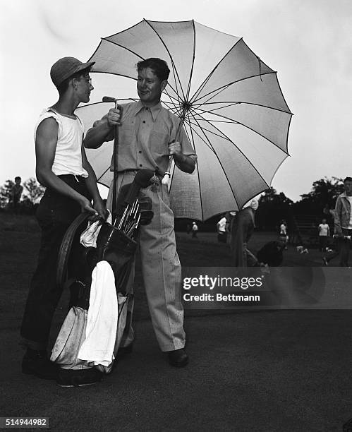 With one hand Byron Nelson takes putter from bag held by caddy Donnie Fox, and with the other he holds a big umbrella to keep off the rain which...