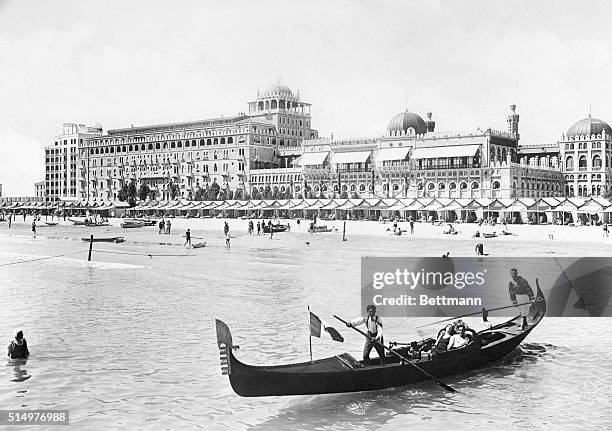 General view of the Excelsior Palace Hotel and lido in Venice, Italy. A gondallier rows two tourists in front of hotel. Undated photograph.