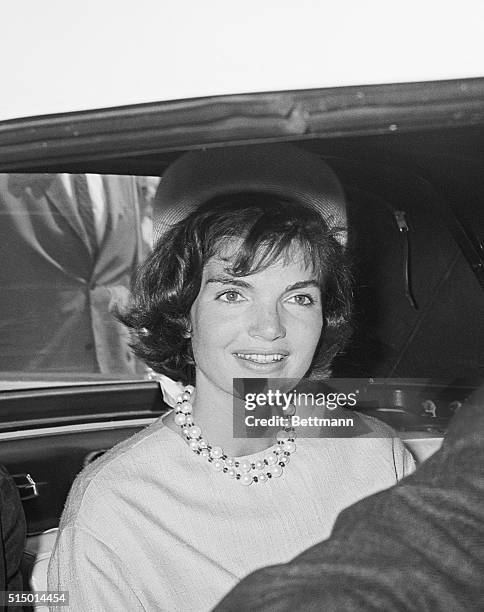 Wearing a straw pillbox hat and a pale blue silk dress, Mrs. Jacqueline Kennedy smiles from the rear seat of an auto as she and the President head...