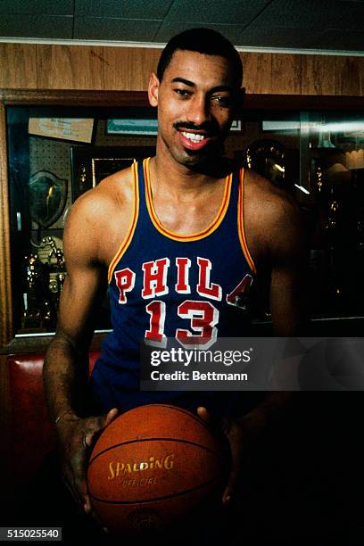 Philadelphia Warriors basketball player Wilt Chamberlain holds a basketball while leaning against a trophy case.