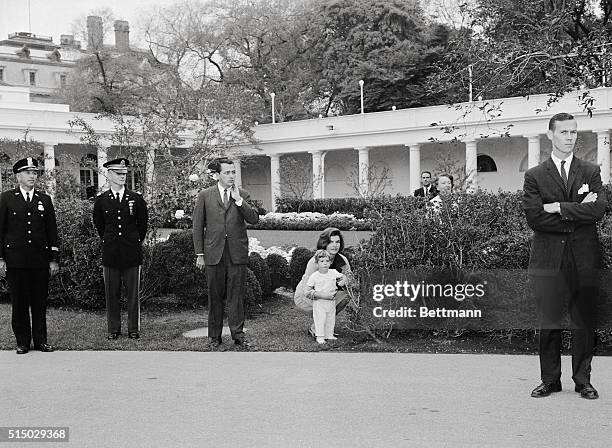 Trying to stay in the background, Mrs. Jacqueline Kennedy and her son, John Jr., peep out from behind shrubbery as they watch ceremonies on the south...