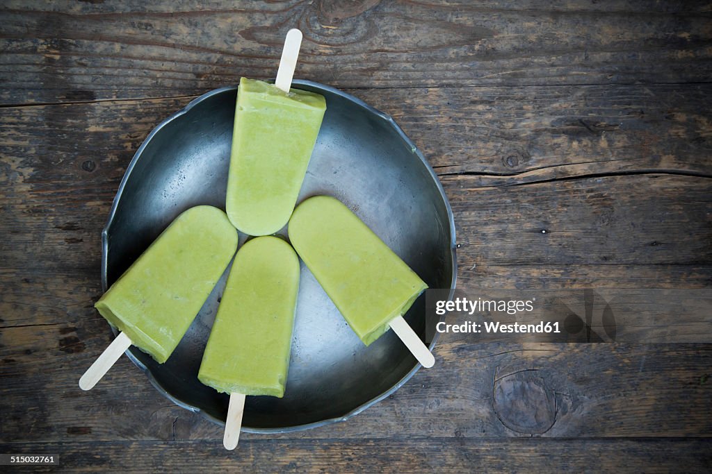 Bowl with four avocado ice lollies on dark wood, elevated view