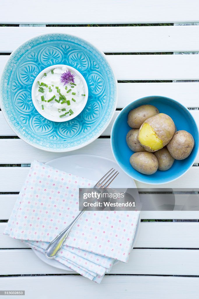 Bowl of jacket potatoes and bowl of curd with herbs, cloth and fork on white wooden table