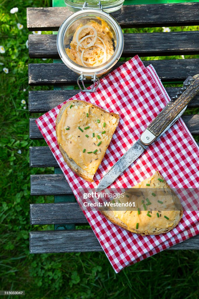 Two halves of slice of bread with obazda sprinkled with chives, cloth, knife and preserving jar on garden table