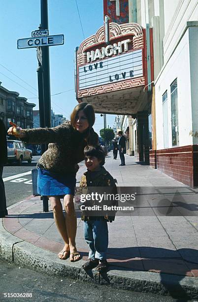 Female hippie and young boy stand on a street corner in the Haight Ashbury area. Theater marquee in background displays the message "Love."