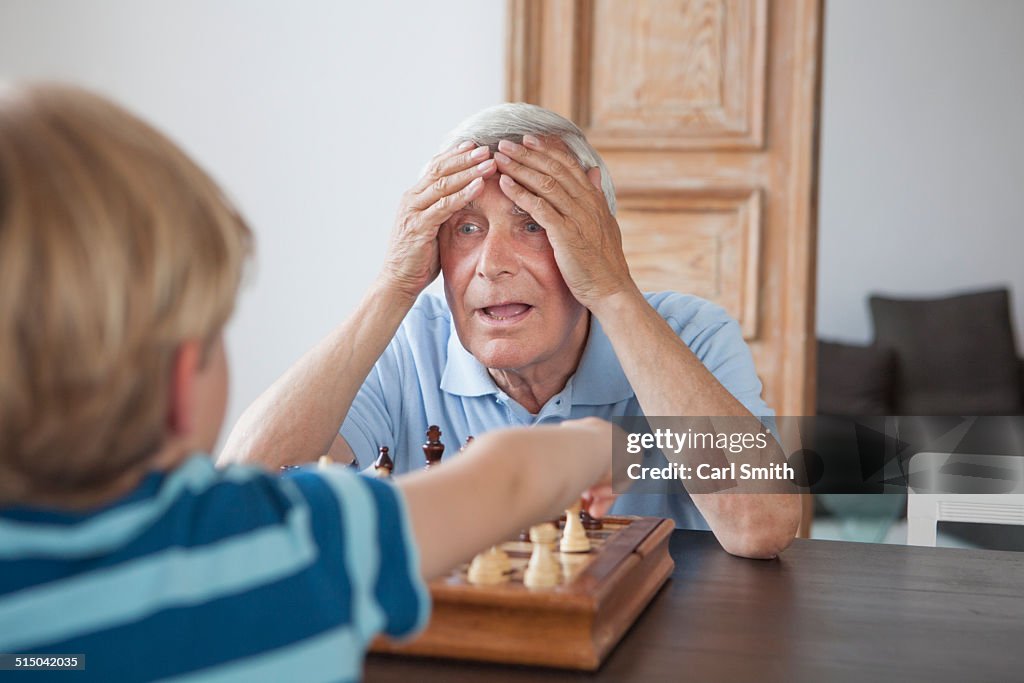 Shocked senior man with head in hands looking at grandson while playing chess at home