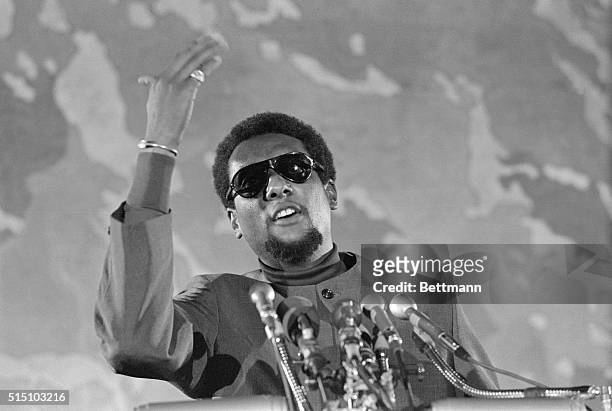 This photograph shows Stokely Carmichael wearing sunglasses as he speaks passionately before a bank of microphones.