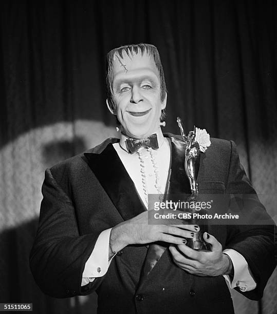 American actor and children's book author Fred Gwynne as 'Herman Munster' stands in a tuxedo before a curtain with an award trophy in a still from...