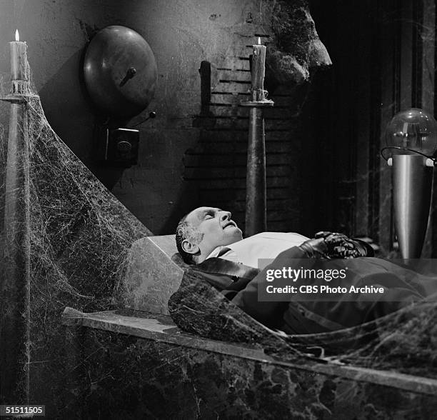 American actor Al Lewis in the role of 'Grandpa' wears evening dress as he sleeps in a cobwebbed room beneath a large alarm bell in a still from the...