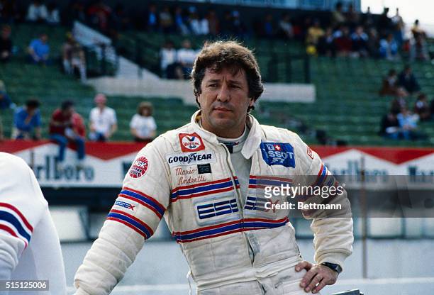 Close-up shows auto racer Mario Andretti at the Spanish Grand Prix. Here, one hand is on one of his hips while another rests on a waist-high object.