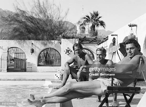 Johnny Weismuller , Bill Acton , swimming pro; and "Stubby" Krueger, prominent Olympic diver, sun themselves beside the pool at the El Mirador Hotel...