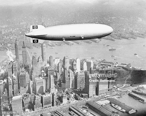 The Hindenburg flies over lower Manhattan on her maiden transatlantic flight. Past the tail of the zeppelin can be seen the Bank of Manhattan...