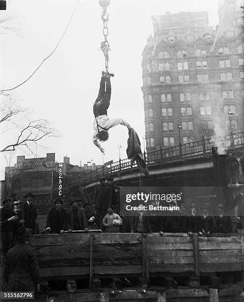 Spectators watch as escape artist Harry Houdini frees himself from a straightjacket while he hangs from a hook above a subway.