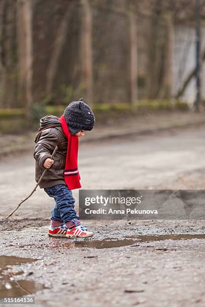 toddler boy with a stick playing ina puddle - eenjarig plantenkenmerk stockfoto's en -beelden