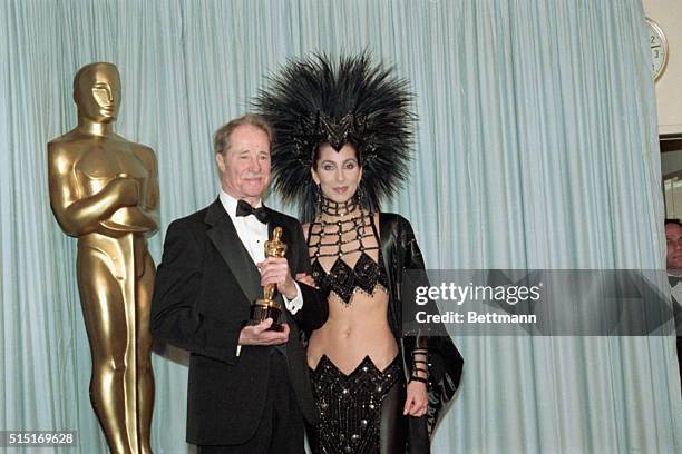 Don Ameche holding the Best Supporting Actor Oscar award for his role in Cocoon, backstage at the Academy Awards with award presenter Cher, who is...