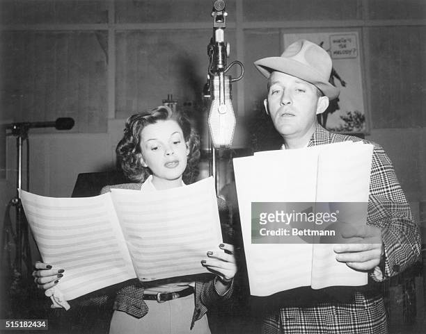 Photograph of Judy Garland and Bing Crosby singing into a microphone in a studio, reading from sheet music. Crosby wears a hat. Undated photograph.
