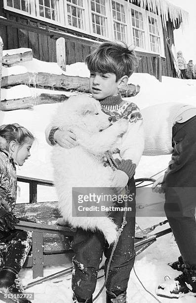 John F. Kennedy Jr. Holds a fuzzy samoyed puppy as his sister Caroline and his mother, Jacqueline Kennedy Onassis, move a dogsled. The family was...