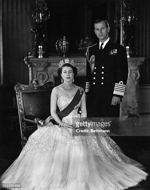 Queen Elizabeth II and Prince Phillip. She is seated and wearing a crown, he is standing in uniform. Undated photograph.