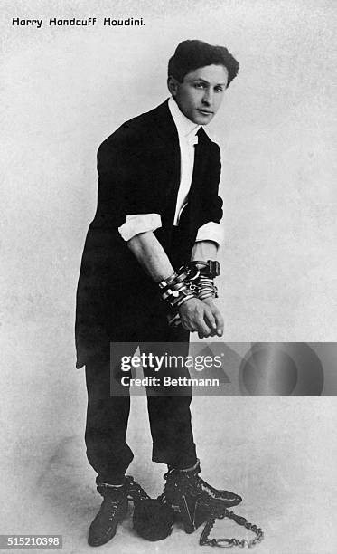 Portrait of American escape artist Harry Houdini dressed in a tuxedo and bound by ankle shakles and wrist handcuffs. Undated photograph.