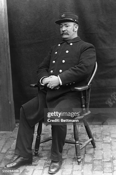 Fire Chief poses for photograph. He sits in a chair on a brick sidewalk, turned at a 3/4 position. He wears a uniform appropriate to his station,...