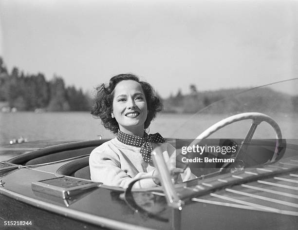 Merle Oberon, on location for her latest picture, Night Song, sits behind the wheel of a motorboat.