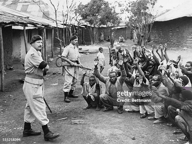 British policemen hold men from the village of Kariobangi at gunpoint while their huts are searched for evidence that they participated in the Mau...