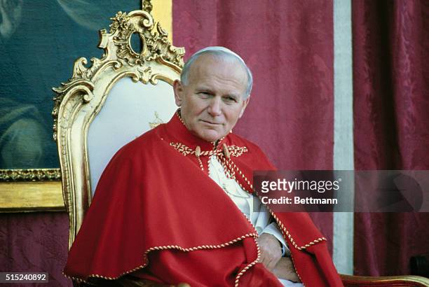 Avila, Spain: Close-up of Pope John Paul II sitting on a chair, wearing red robes with a portrait of the St. Theresa of Avila in the background...