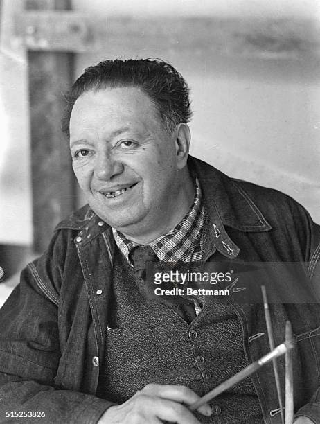 This is a waist-up photo of Mexican Diego Rivera, , smiling as he reaches into an inner pocket of his jacket.