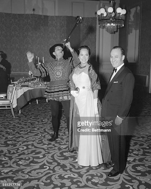 Actress Merle Oberon and husband Bruno Pagliai, a Mexican businessman, get loud greeting from a costumed trumpeter as they arrive for a society...