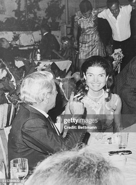 Jackie Onassis smiles at photographer while her husband, Aristotle, admires the "syrtake" skill of some of his friends dancing , at a party...