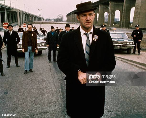 Policeman Gene Hackman is shown in this movie still walking toward the camera. He is holding a gun in his hands with policemen and police vehicles in...