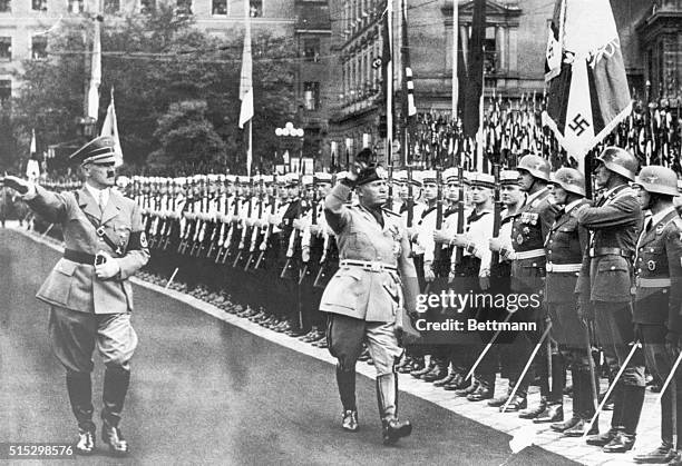 Adolf Hitler with Benito Mussolini reviewing Nazi troops. They are saluting as they walk down the orderly line of soldiers.