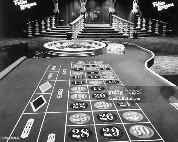 Photo is a view looking down a roulette table towards the doors of a casino, with signs on either side of the door that read "Folies Bergere."...