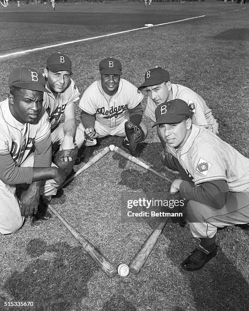 Gathered around a miniature diamond in approximately the same spots they would cover on the big one, are five members of the Brooklyn Dodgers. Left...