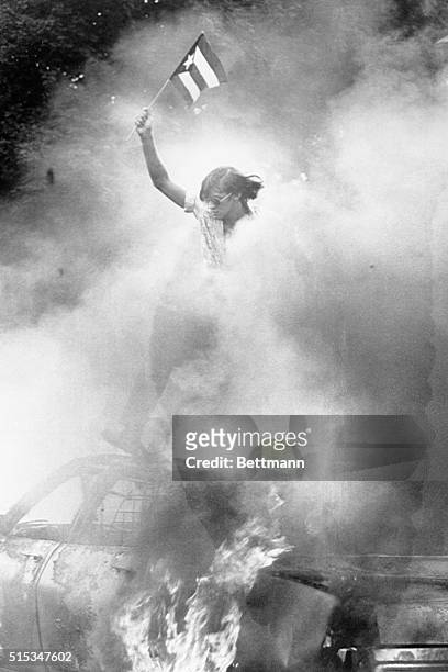 While a Newark Plice Department patrol car burns, a young Puerto Rican holds a small flag high over the wreckage near Branch Brook Park as a Puerto...