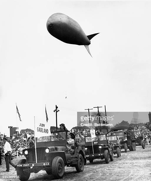 Beneath a barrage balloon, Judy Garland, screen star, joins the Jeep parade of Hollywood celebrities that opened the Treasury's ‘Back the Attack'...