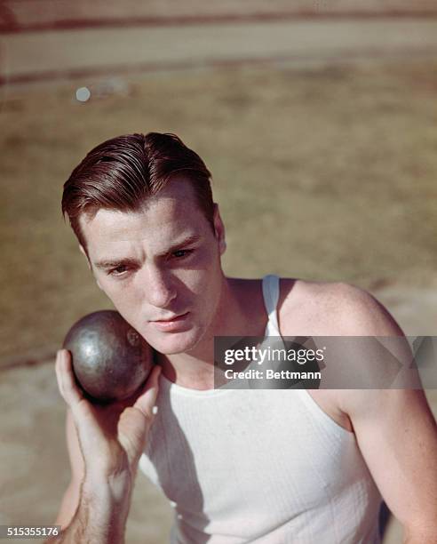 Olympic Gold medalist Bob Mathias is show holding the shot put next to his head.