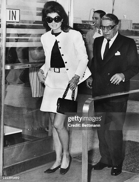 Wearing mod sunglasses and a miniskirt, Jacqueline Kennedy Onassis accompanies her husband, Aristotle, to a plane at Kennedy Airport on today, before...