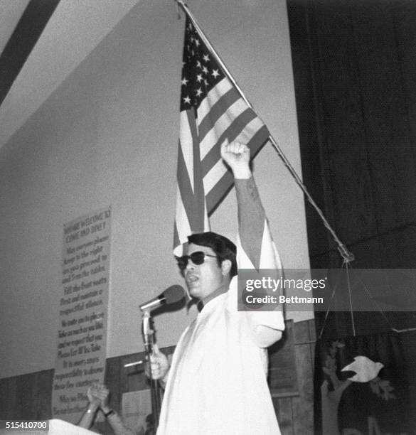 Reverend Jim Jones raises his fist in a black power salute while preaching at an unknown location. This is one of the photographs from a photo album...