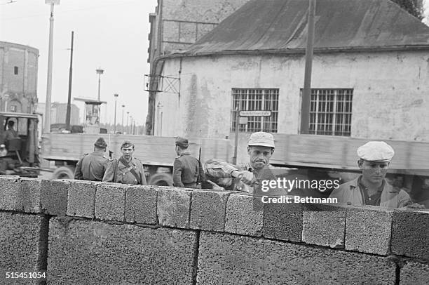 Berlin, Germany- Grim-faced East German workers, under the watchful eyes of the Communist police, erect a high, concrete wall at a sector border near...