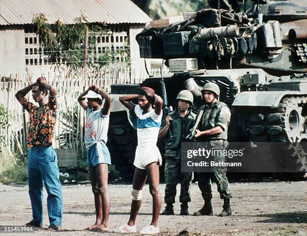 Grenada: A United States combat soldier covers some prisoners of war, who have their hands on their heads in the classic "arrest" pose, during the...